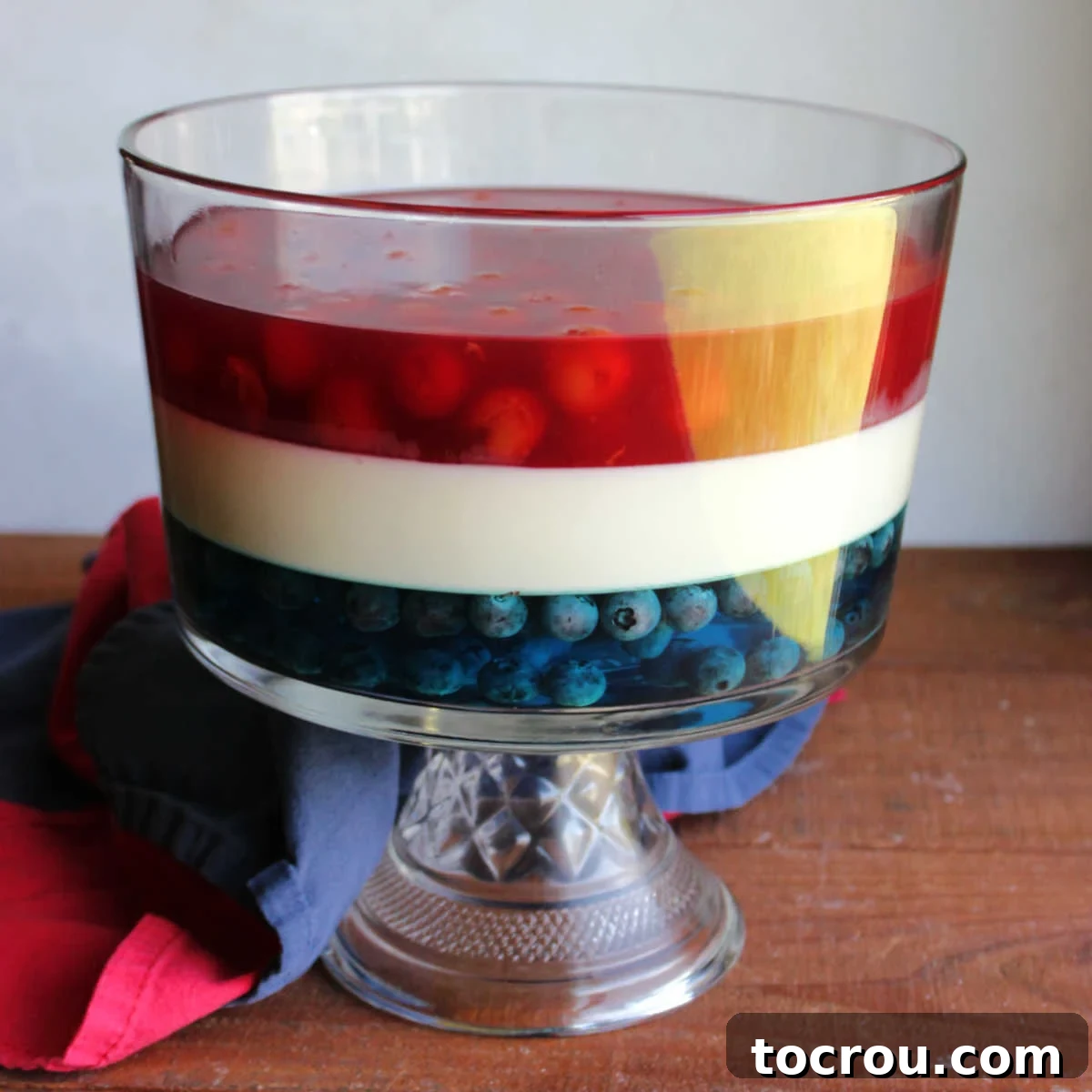 Patriotic Layered Jello 2 A beautiful glass trifle bowl showcasing distinct layers of blue Jello with blueberries, a creamy white Jello layer, and a vibrant red cherry Jello layer, perfect for patriotic parties.