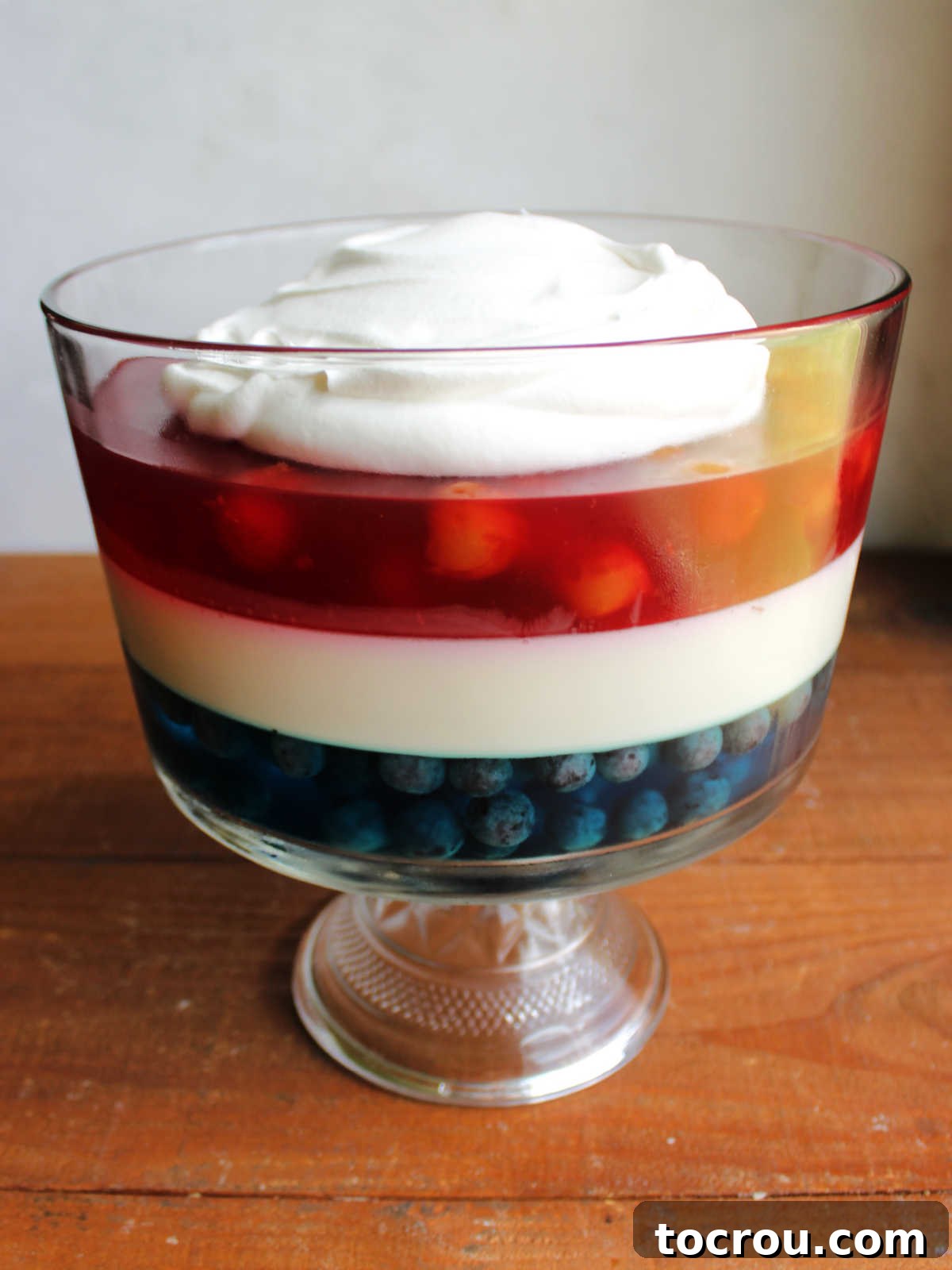 Patriotic Layered Jello 3 Glass trifle bowl showcasing the vibrant layers of blue Jello with blueberries, creamy white Jello, and red Jello with cherries, elegantly topped with whipped cream.