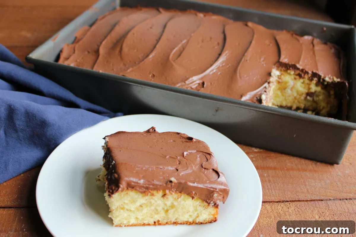 A slice of yellow cake lavishly topped with smooth, rich chocolate Russian buttercream frosting, with the remainder of the cake and more frosting visible in the background, inviting a delicious bite.
