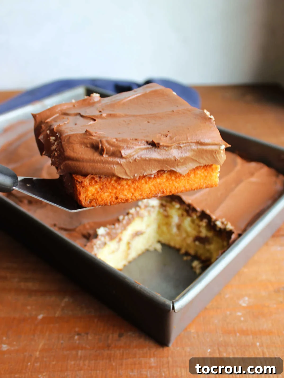 A spatula gently lifts a perfectly frosted slice of yellow cake, adorned with smooth Russian chocolate frosting, from a 9x13-inch baking pan, highlighting its inviting texture.