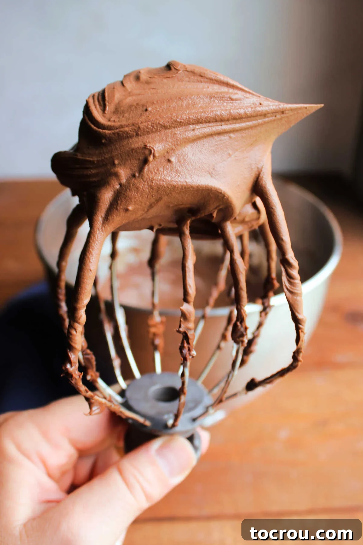 A hand holds a mixer beater, richly coated with fluffy, light chocolate Russian buttercream, ready to frost a delicious cake or batch of cupcakes.