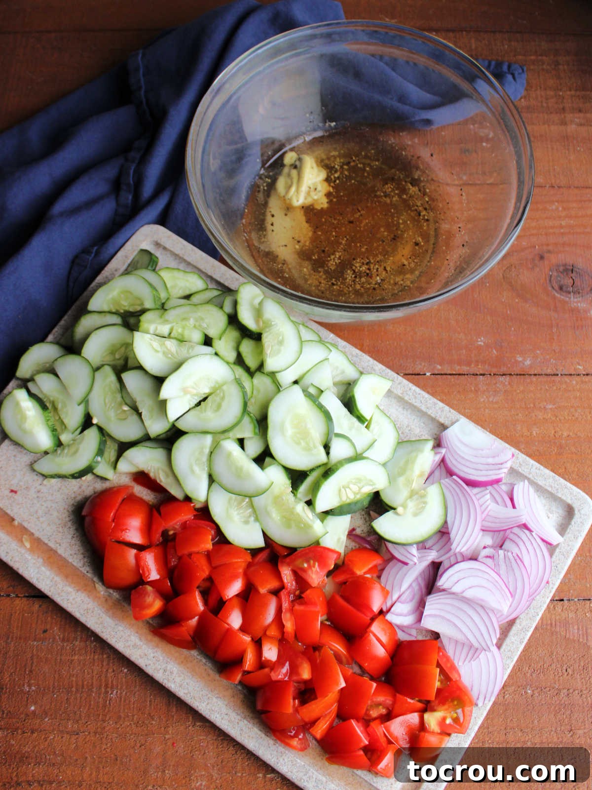 Ingredients for fresh cucumber salad: sliced cucumbers, chopped tomatoes, and sliced red onion, next to a bowl of homemade vinaigrette dressing ingredients including vinegar, oil, honey, salt, pepper, and dijon mustard.