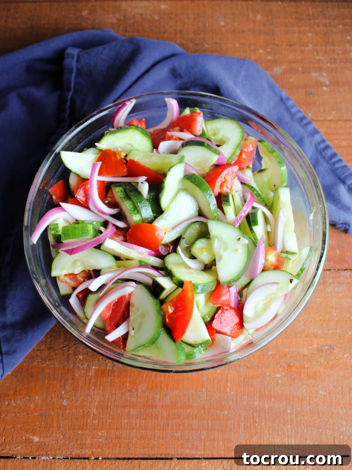 Mixing bowl with fresh cucumber, tomato, and red onion vegetables tossed thoroughly in a homemade vinaigrette, ready to be chilled for marinating before serving.