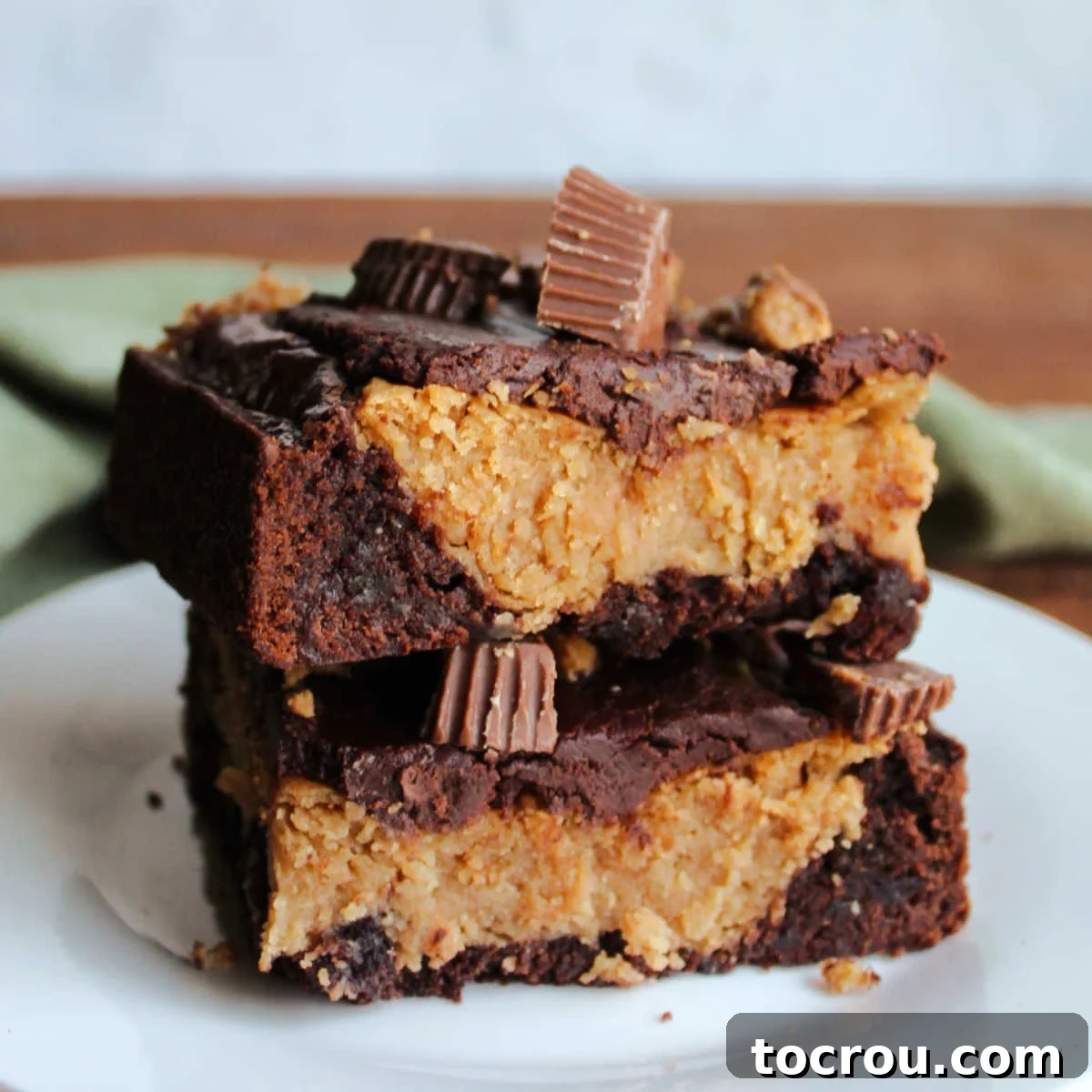 Two square pieces of rich peanut butter cheesecake brownies stacked on top of each other, showcasing the fudgy brownie base, creamy peanut butter cheesecake filling, glossy chocolate ganache, and peanut butter cup toppings.