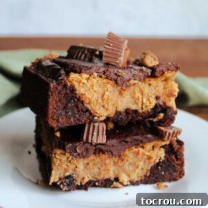 Two square pieces of peanut butter cheesecake brownie stacked on top of each other showing brownie base, rich peanut butter cheesecake filling and ganache and peanut butter cups on top.