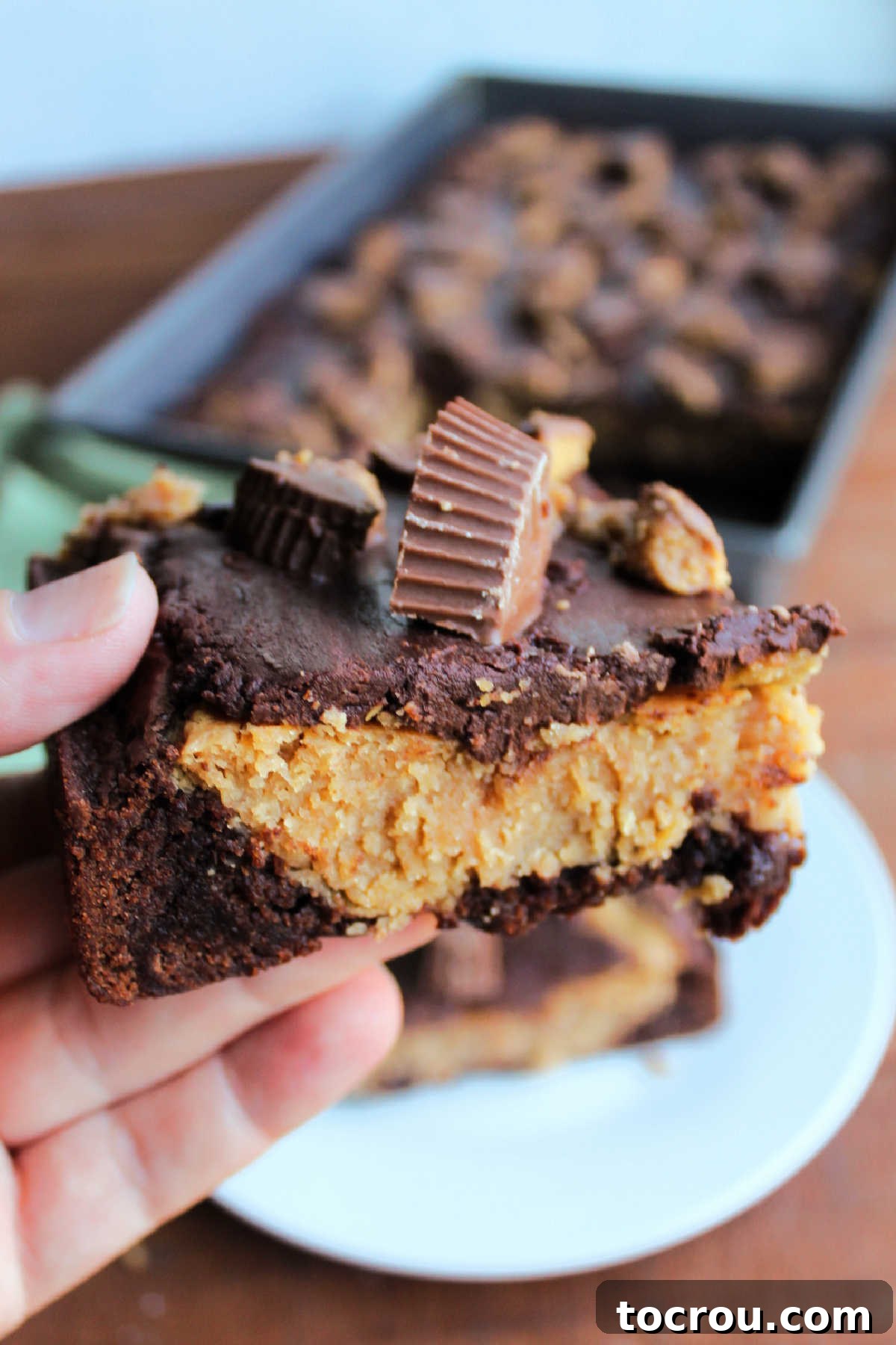 A hand holds a square of peanut butter cheesecake brownie, showcasing its distinct layers of brownie, peanut butter cheesecake, and chocolate ganache with peanut butter cup pieces, ready for a bite.