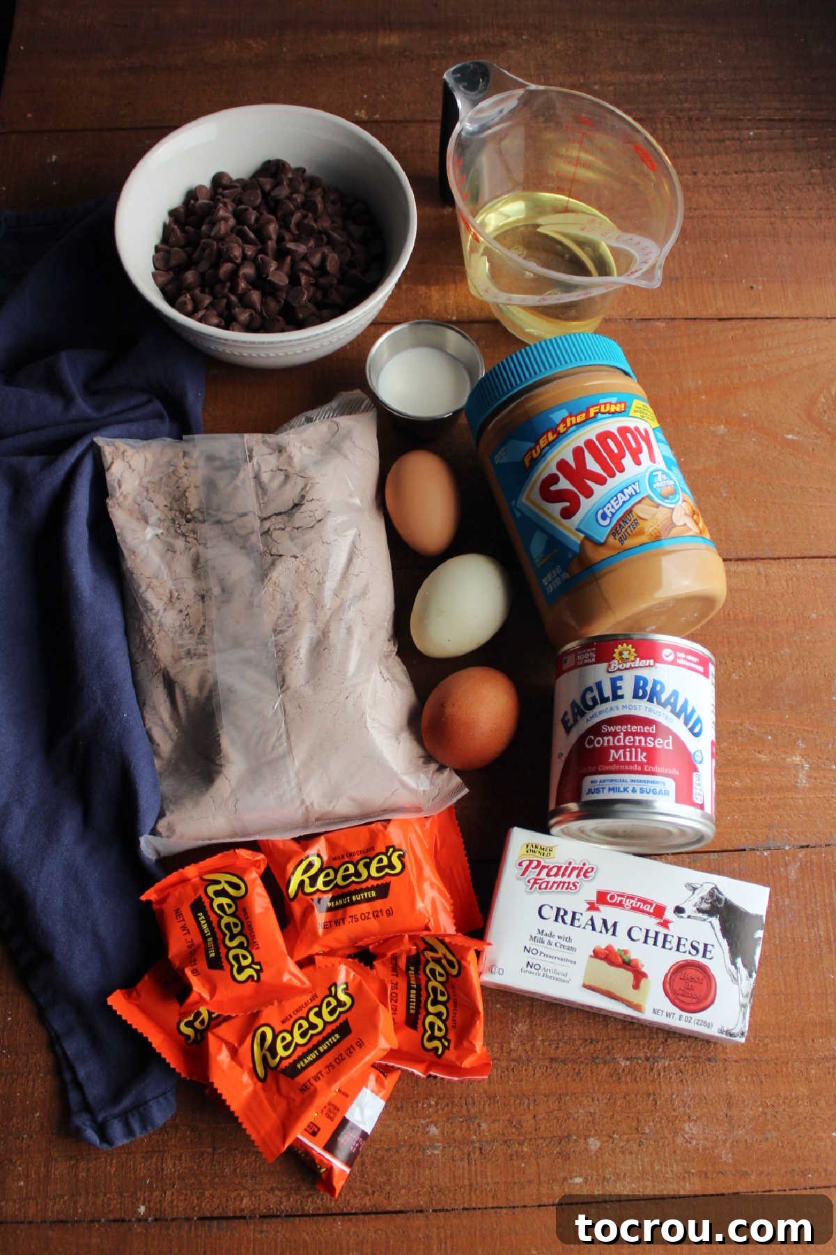 A selection of ingredients laid out on a kitchen counter, including a brownie mix box, eggs, a jar of peanut butter, oil, a can of sweetened condensed milk, cream cheese, chocolate chips, and Reese's peanut butter cups, all prepared for making peanut butter cheesecake brownies.