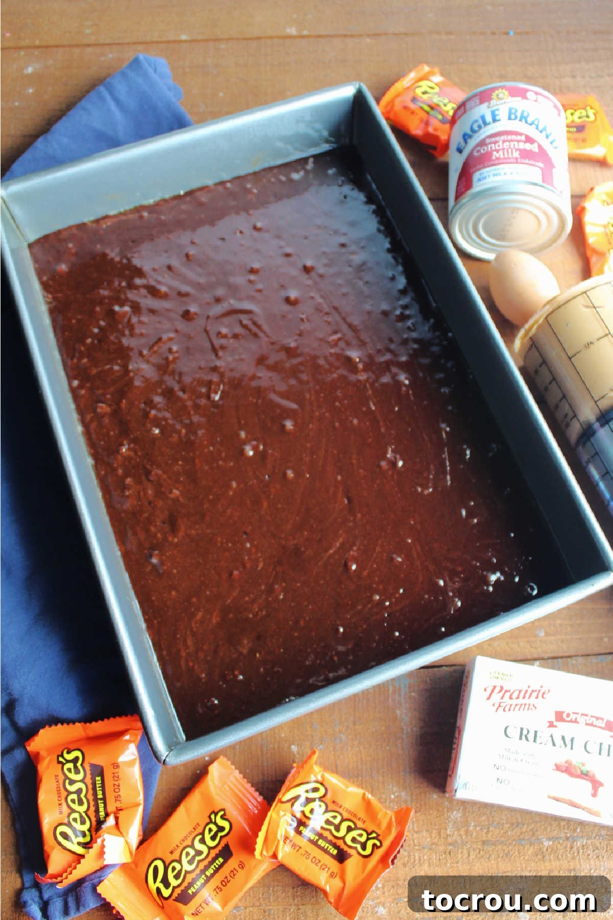 Smooth brownie batter spread evenly in a 9x13-inch baking pan, with additional ingredients like cream cheese, peanut butter, and sweetened condensed milk waiting to be mixed nearby.