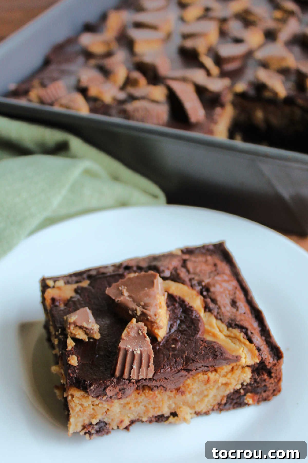 A single piece of peanut butter cheesecake brownie sits on a white plate, showcasing its delightful layers and tempting chocolate and peanut butter cup topping, ready to be devoured.