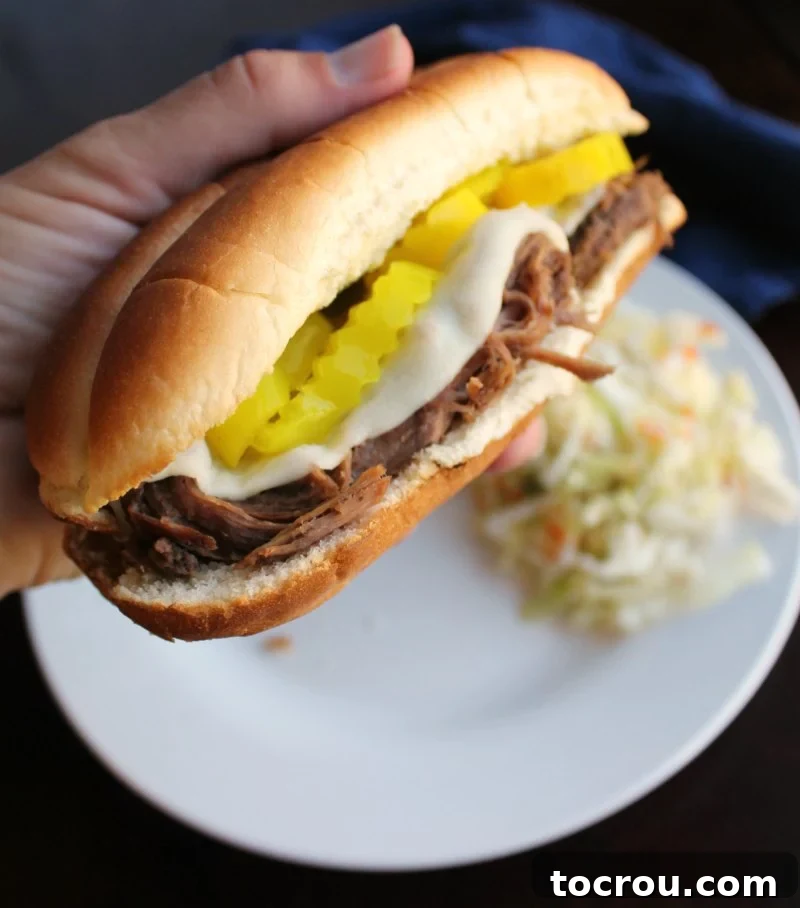 Hand holding ang Italian beef sandwich on a hoagie roll with melted cheese and peppers on top.