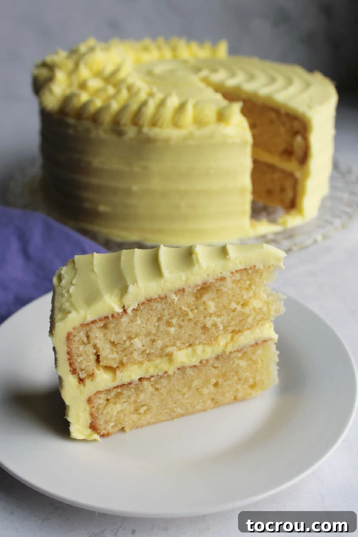 Lemon Velvet Cake Slice A perfect slice of moist lemon velvet cake, beautifully frosted with a generous layer of lemon condensed milk buttercream, rests invitingly on a white dessert plate, with the remainder of the elegant cake visible in the softly blurred background.