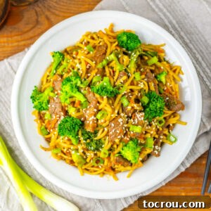 Beef and Broccoli Lo Mein Dinner plate filled with lo mein noodles with bits of steak and broccoli in garlic soy sauce topped with sesame seeds and scallions, ready to eat.