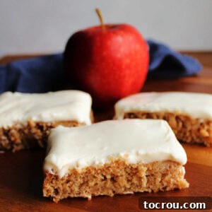 Individual pieces of moist applesauce cake topped with soft white sour cream frosting, with a fresh apple blurred in the background, suggesting autumnal flavors.