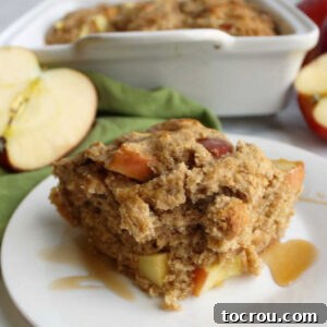 A close-up of a slice of rustic buttermilk apple cake, with apple halves and a cake pan visible in the soft background, highlighting its homemade charm.