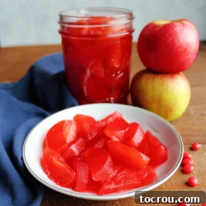 A close-up of a small bowl filled with bright red stewed apple slices, positioned next to a jar of candied apples, creating a vivid display of cinnamon-infused treats.