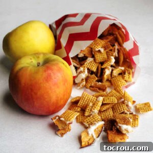 Two fresh apples sitting next to a bag of homemade apple cinnamon Chex mix, featuring chunks of dried apples and a sweet white chocolate drizzle.