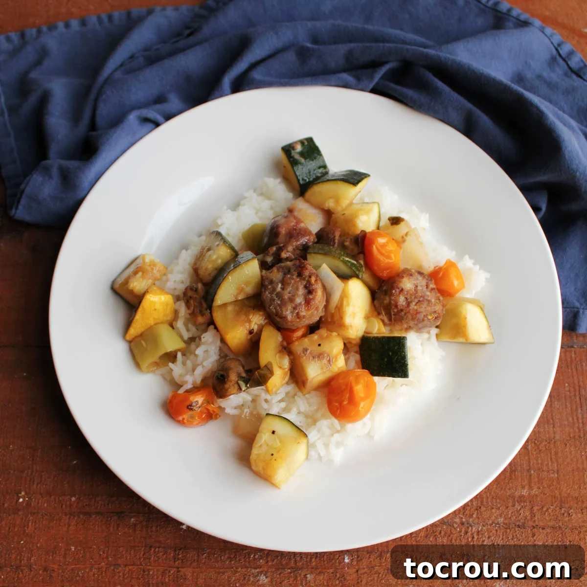 One-Pan Italian Sausage and Roasted Vegetables 2 Dinner plate with rice under roasted Italian sausage chunks and vegetables including zucchini, yellow squash, cherry tomatoes, and more.