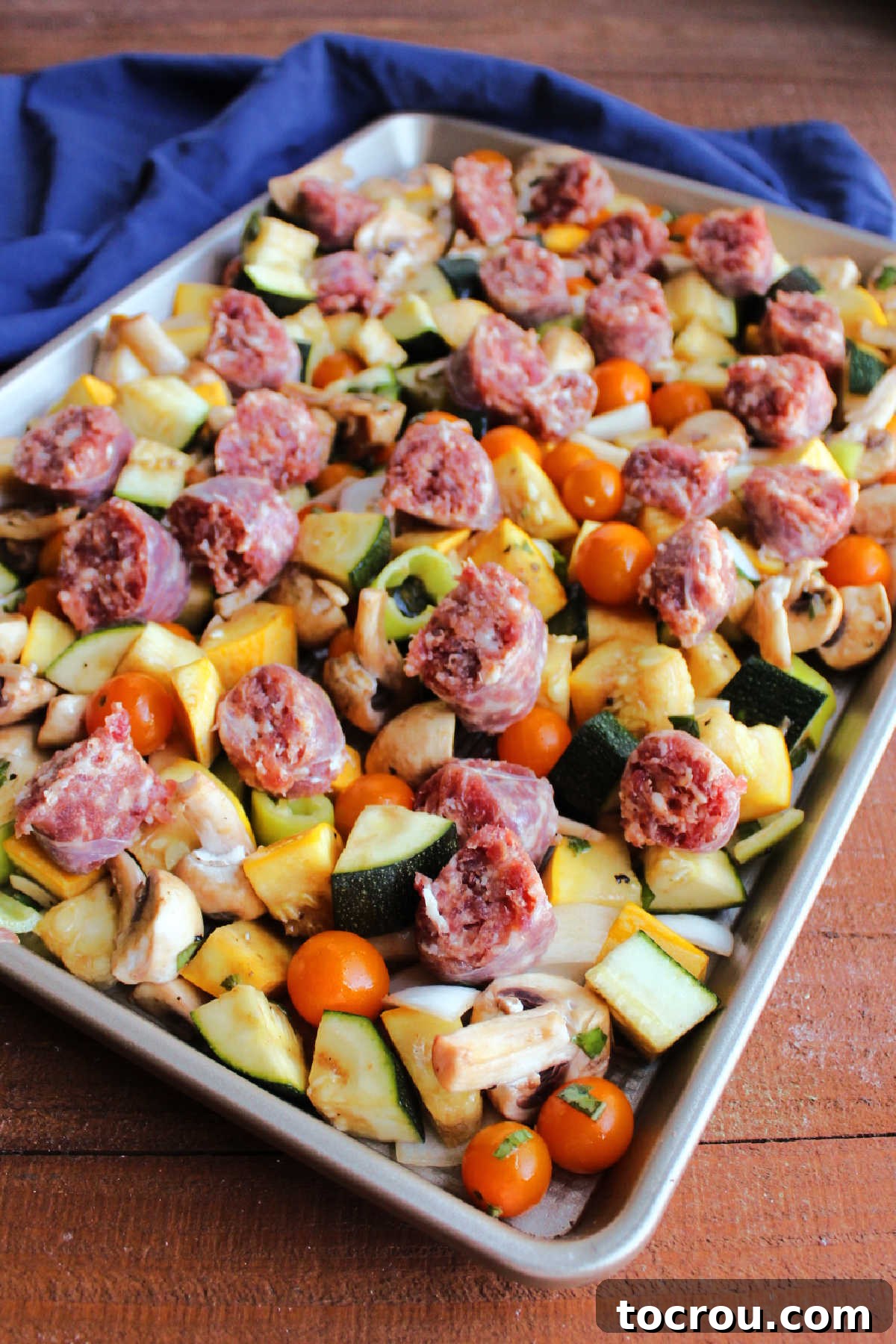 One-Pan Italian Sausage and Roasted Vegetables 7 Vegetables on rimmed sheet pan topped with pieces of raw Italian sausage links, ready to be roasted.