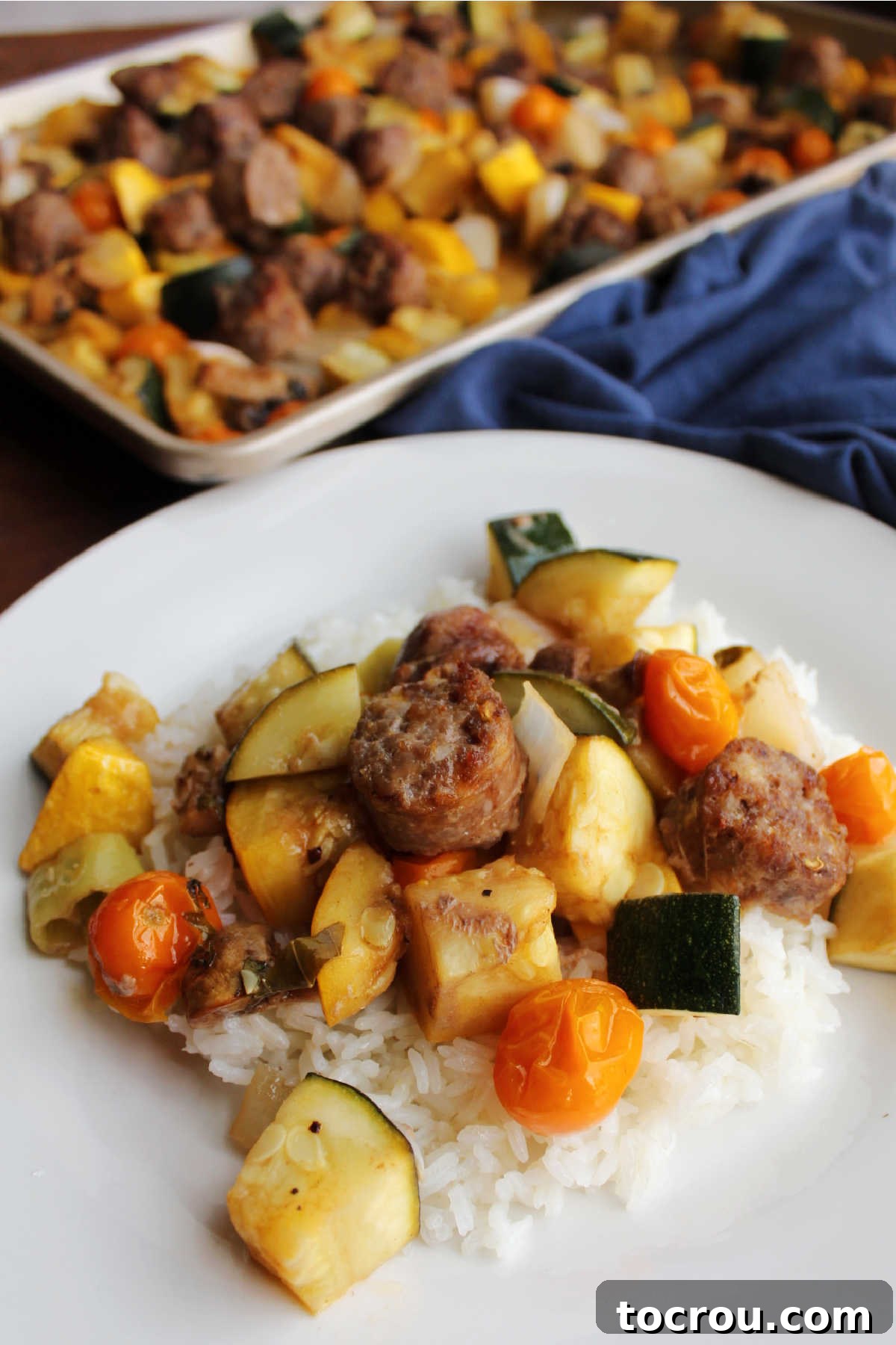 One-Pan Italian Sausage and Roasted Vegetables 9 Close up of Italian sausage and vegetables served over rice with sheet pan of remaining sausage and veggies in the background.