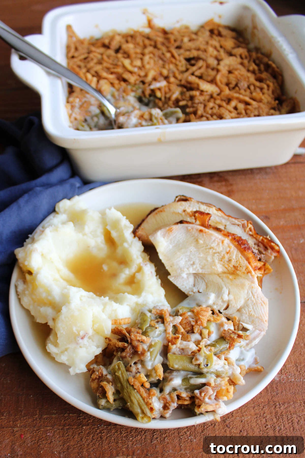 A beautifully plated dinner featuring classic green bean casserole, creamy mashed potatoes with rich gravy, and perfectly sliced roasted turkey breast, set against a backdrop of a full casserole dish.