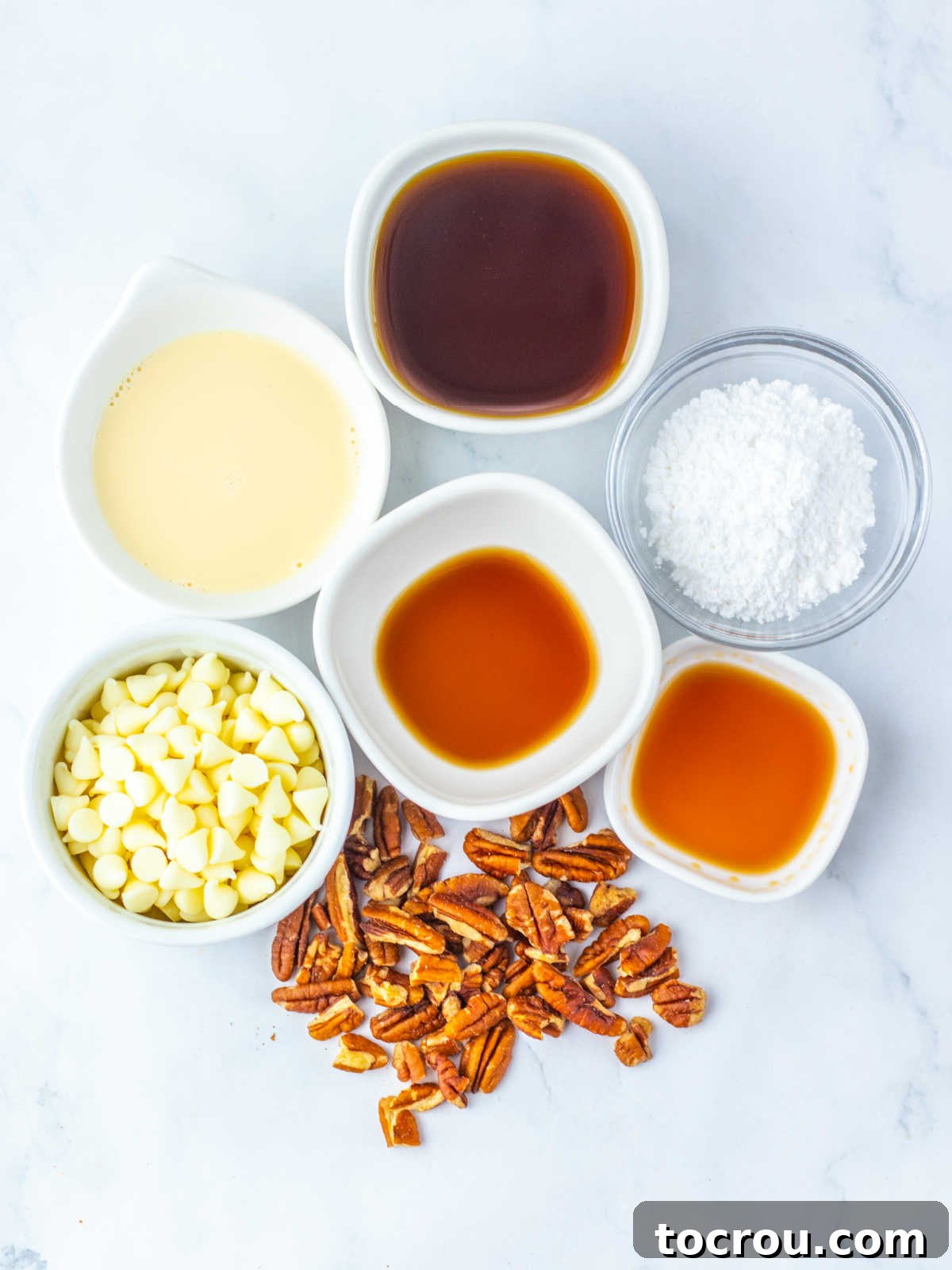 Ingredients including pecans, white chocolate chips, evaporated milk, dark corn syrup, maple extract, vanilla extract, and powdered sugar ready to be made into pecan pie fudge, neatly arranged on a counter.