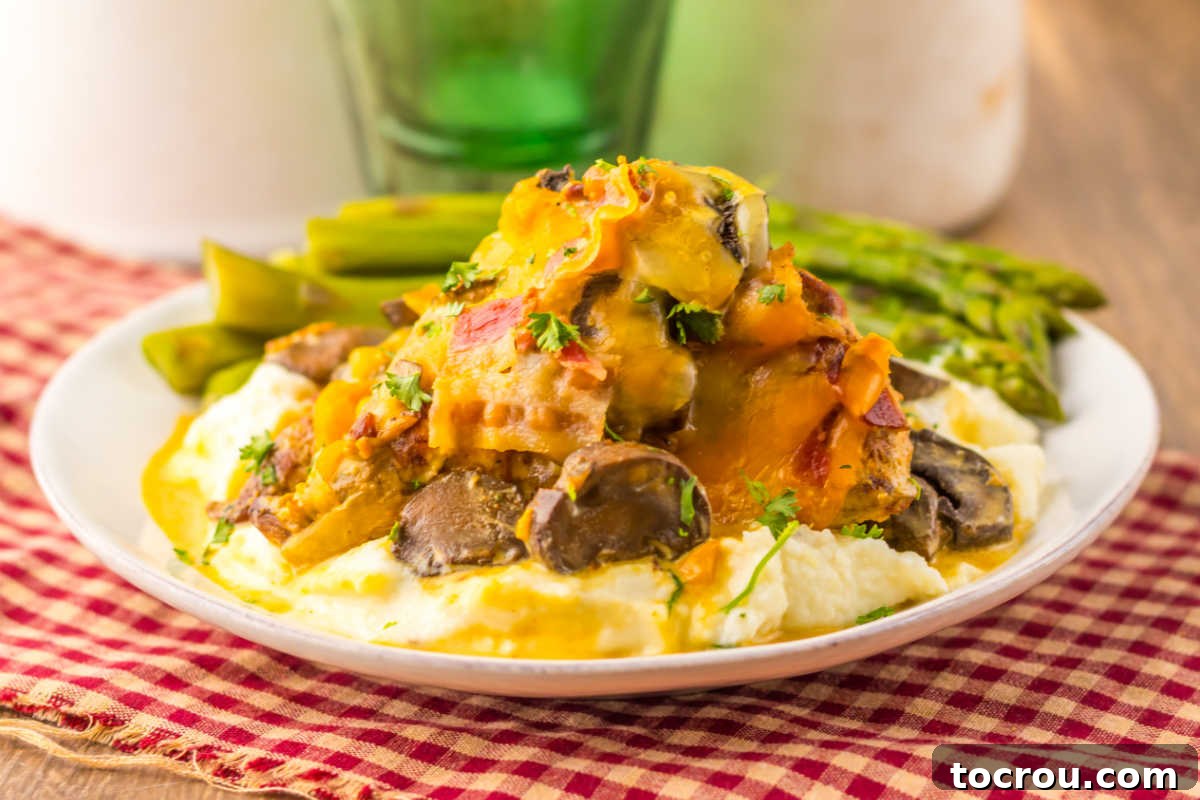 Dinner plate with mashed potatoes topped with a chicken breast with honey mustard sauce, bacon, mushrooms and melted cheese with a side of asparagus in the background.