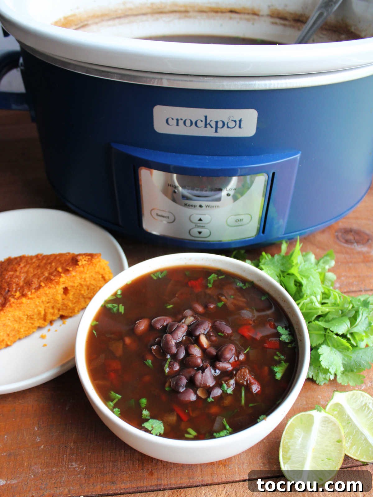 Bowl of black bean soup next to a wedge of cornbread and some cilantro in front of the crockpot it was cooked in.
