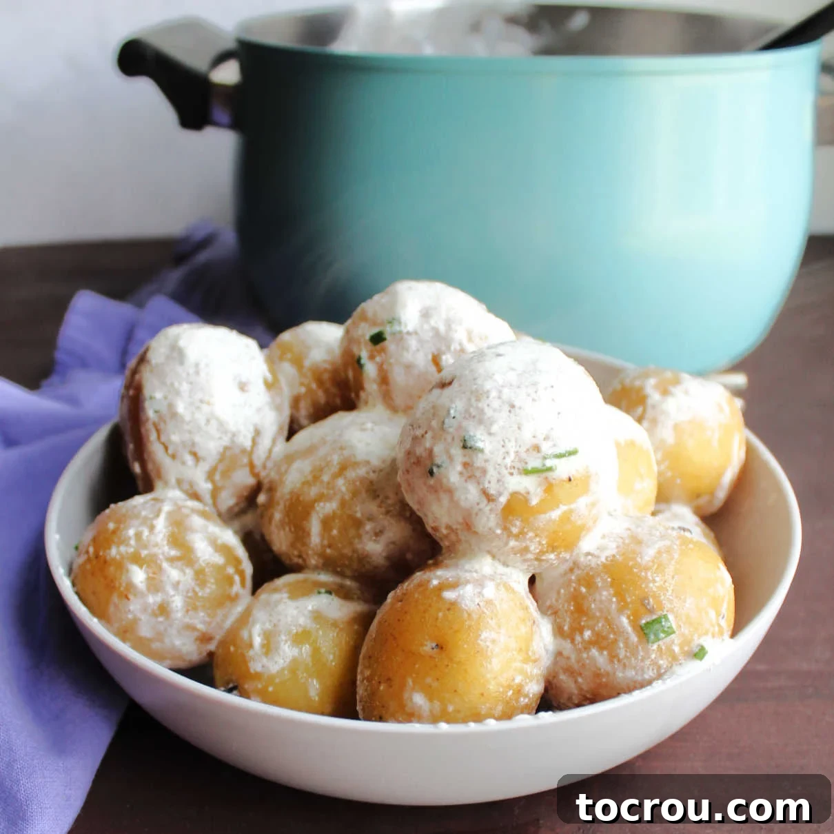 Bowl of new potatoes coated in sour cream and chives in front of pot.