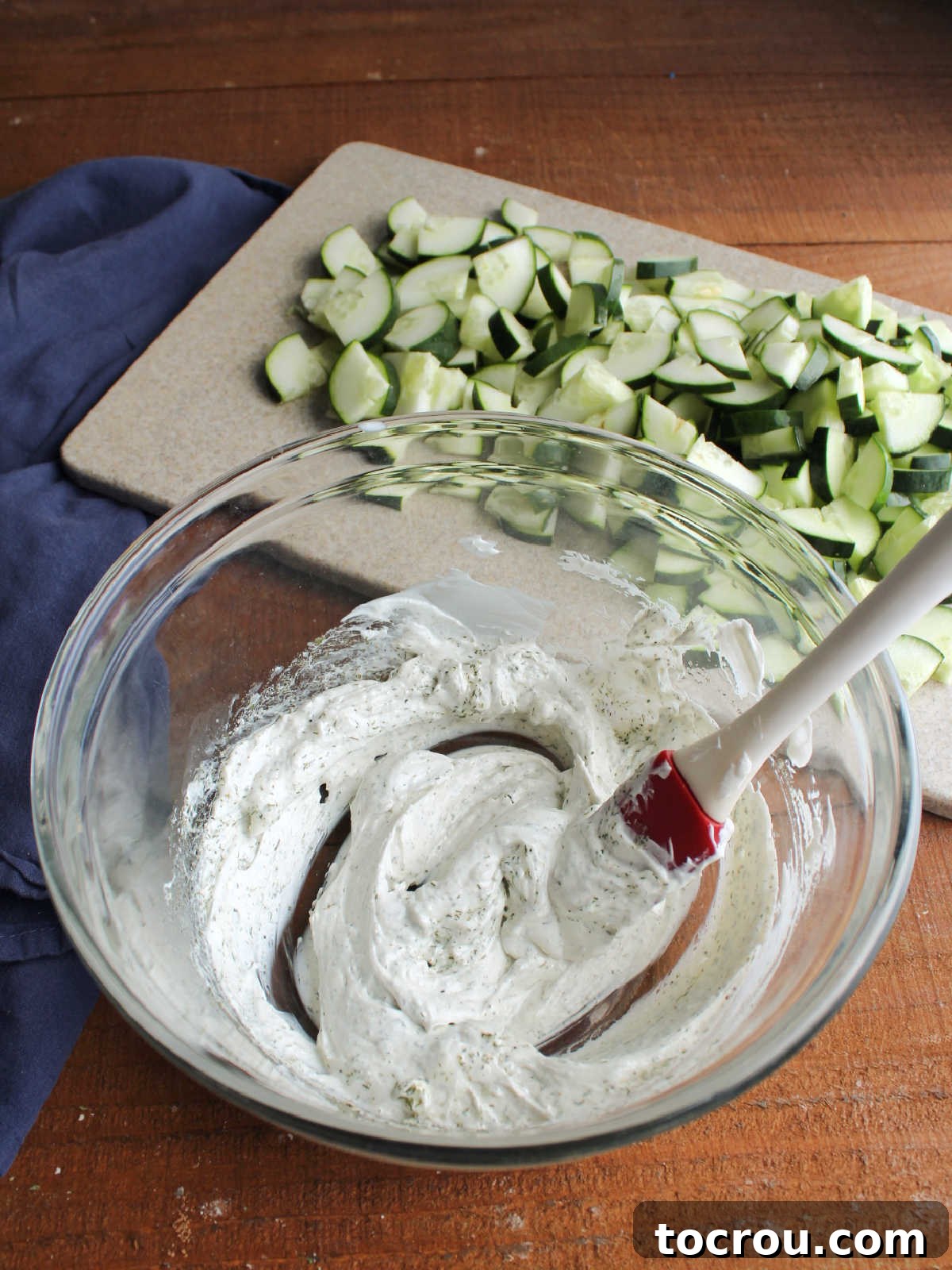 Mixing bowl of sour cream and dill dressing in front of cutting board with cucumber pieces on it.