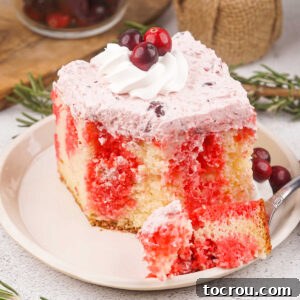 Cranberry Poke Cake A delectable piece of cranberry poke cake, featuring bright red Jello infused into a moist white cake, topped with fluffy cranberry frosting, additional whipped cream, and garnished with fresh cranberries.