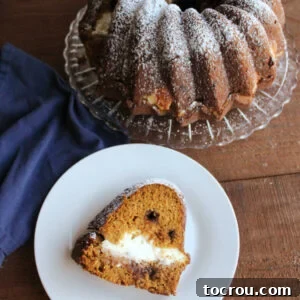 Pumpkin Bundt Cake A slice of moist pumpkin Bundt cake, revealing a creamy cream cheese filling and scattered chocolate chips, is presented on a plate with the remaining powdered sugar-dusted cake in the background.