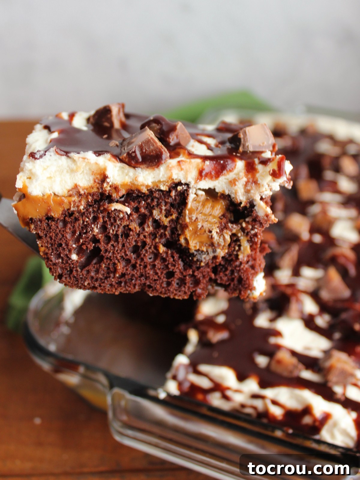 Spatula lifting a generous piece of Rolo poke cake from the pan, clearly showing the distinct layers: the dark chocolate cake, tunnels of dulce de leche, a thick layer of ermine frosting, drizzled hot fudge, and pieces of Rolo candies.