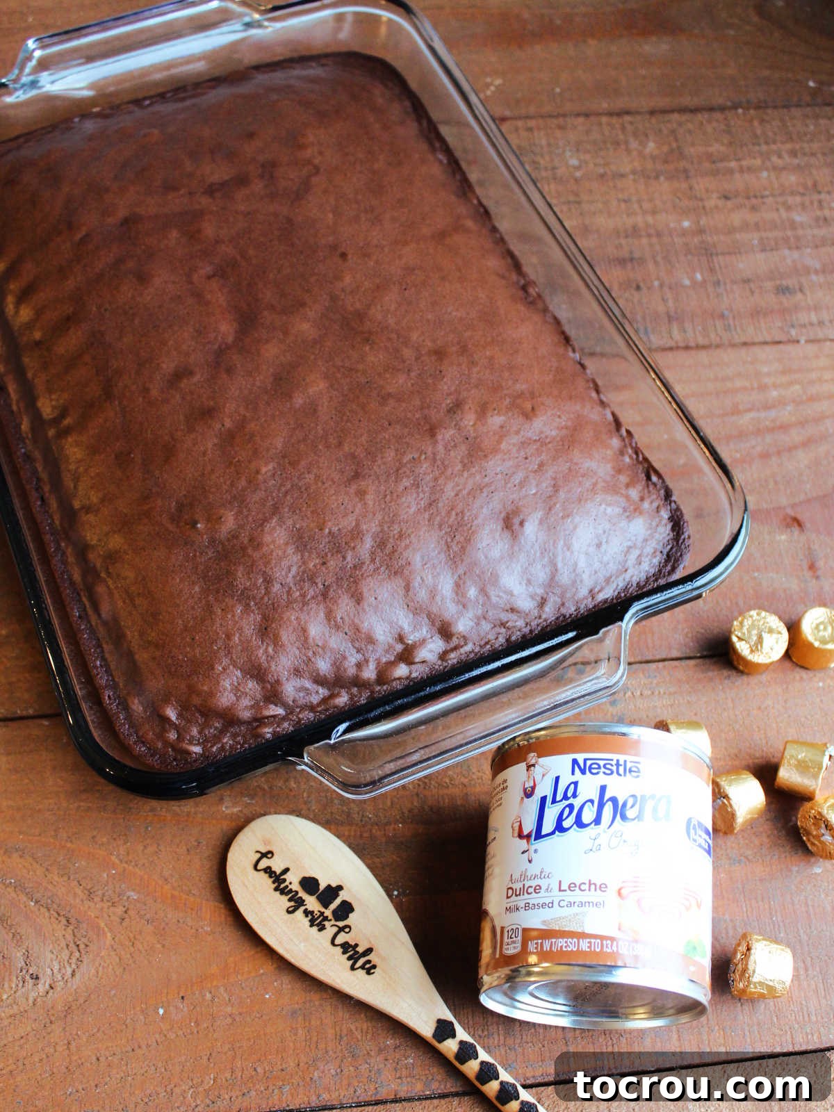 A 9x13-inch pan filled with freshly baked chocolate cake, positioned next to a can of dulce de leche, several Rolo candies, and a wooden spoon, indicating readiness for the next steps in cake preparation.