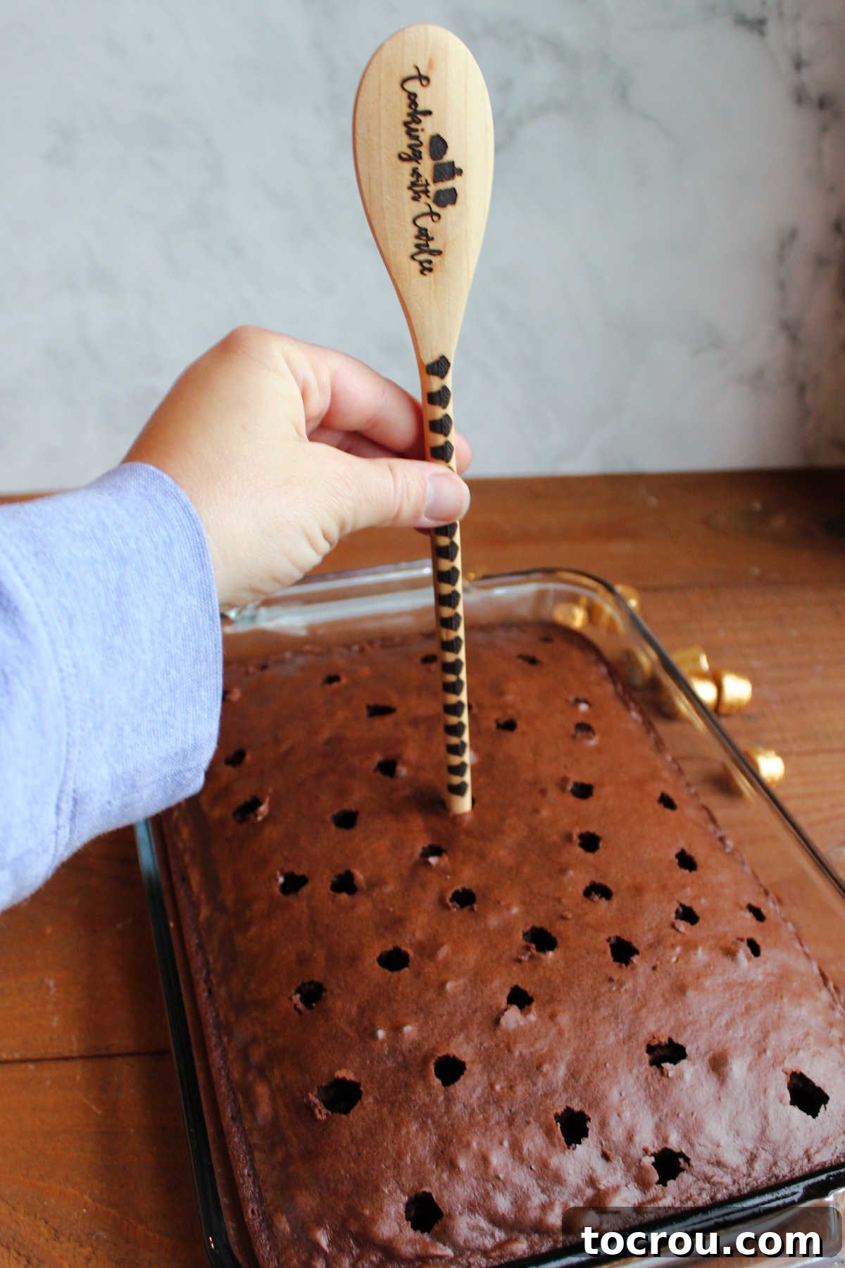 Close-up shot of the process of poking holes evenly across the top of a warm chocolate cake using the round handle of a wooden spoon, preparing it for caramel infusion.