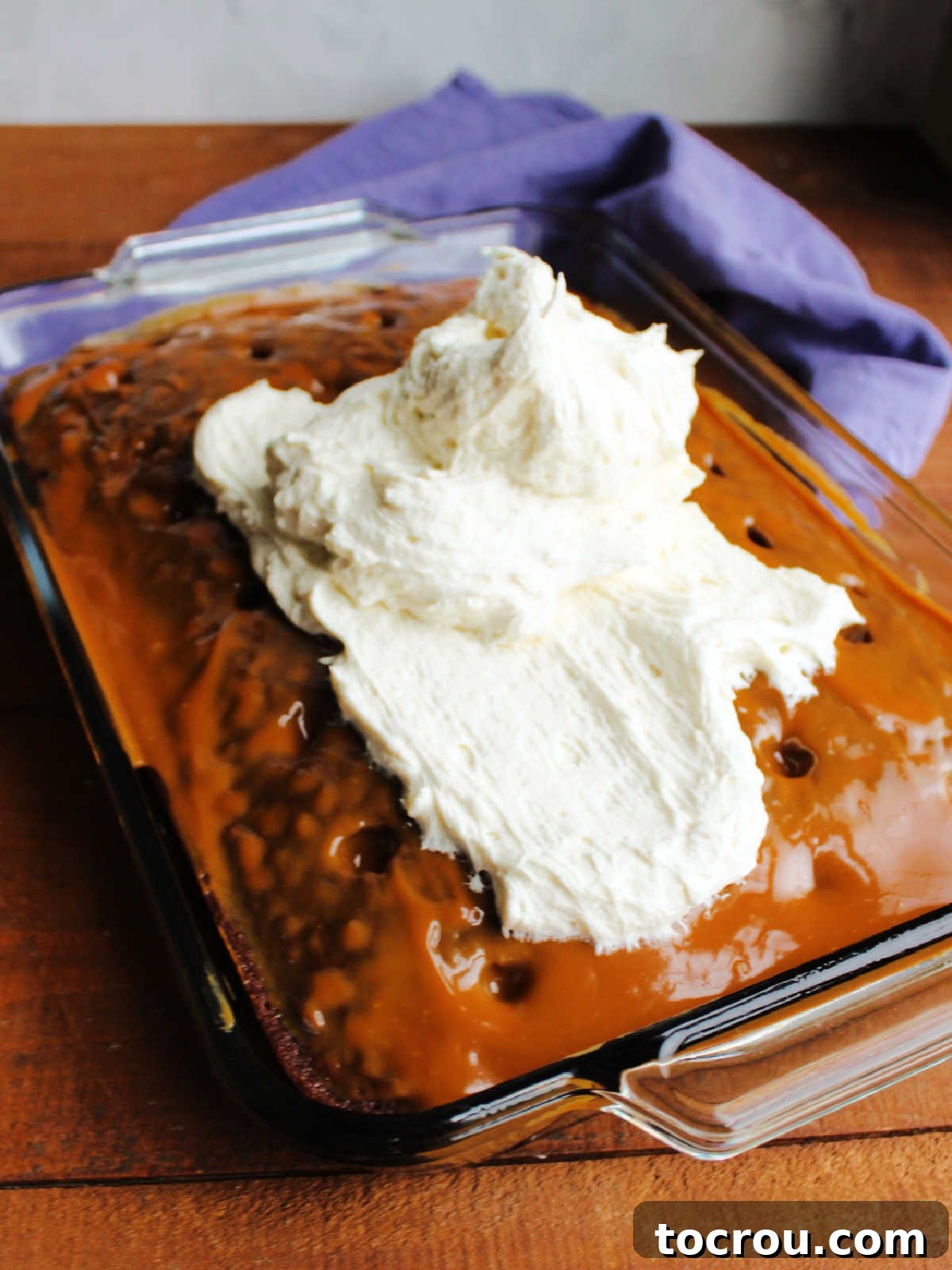 A generous pile of fluffy ermine frosting centrally placed on top of a caramel-coated chocolate cake, awaiting to be spread evenly to create a smooth, inviting layer.