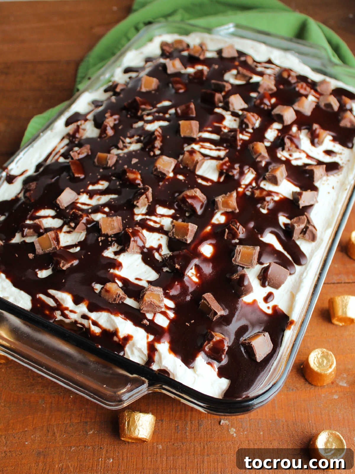 The finished Rolo poke cake, generously topped with fluffy white frosting, an abundant scattering of chopped Rolo candies, and an artistic drizzle of warm hot fudge sauce, ready to be sliced and served.