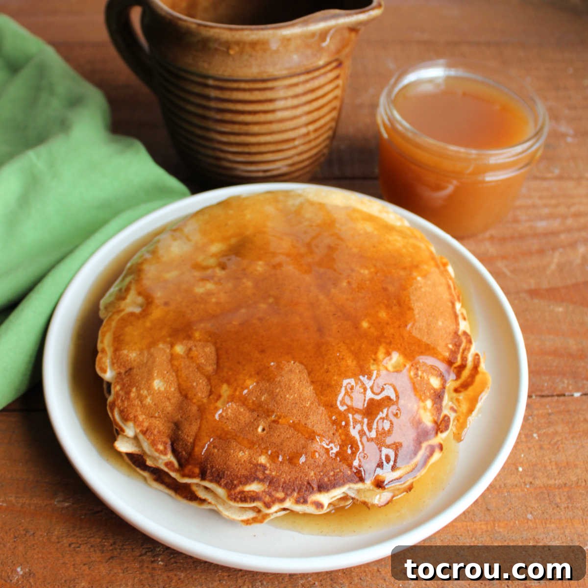 Plate of cinnamon pancakes with apple cider syrup on top with a jar and pitcher of apple cider syrup in the background.