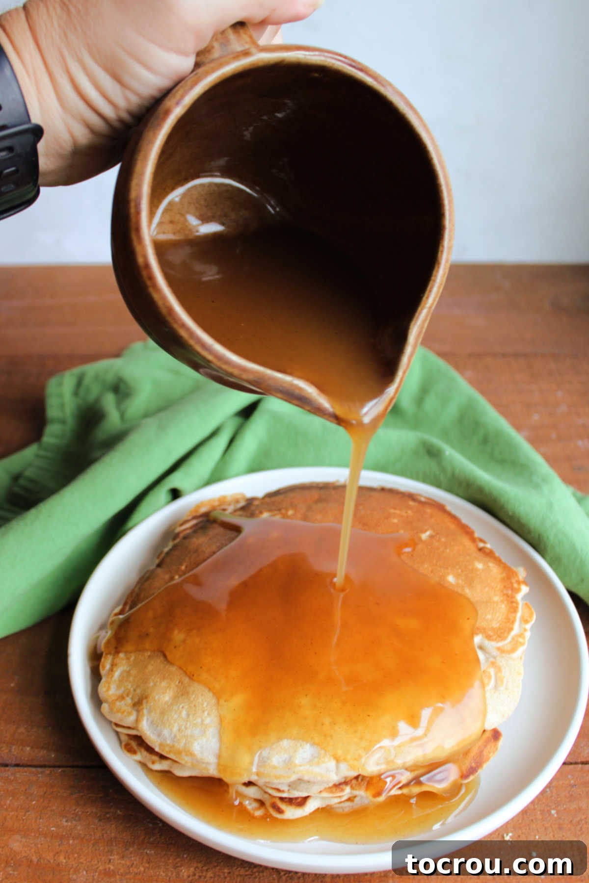 Pitcher of apple cider syrup being poured over a stack of pancakes. 