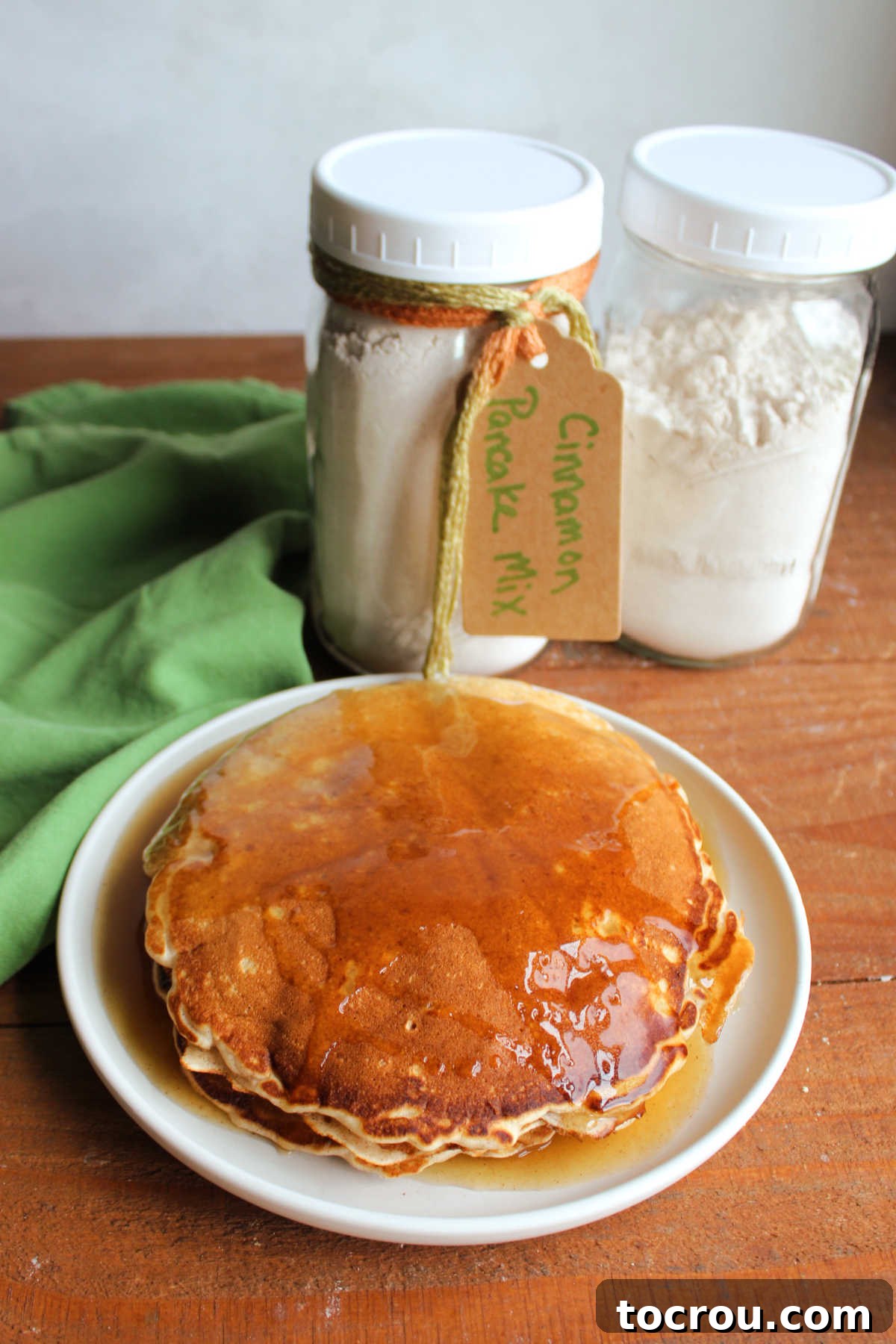 Plate of homemade cinnamon pancakes with apple cider syrup on top next to jars of homemade cinnamon pancake mix.