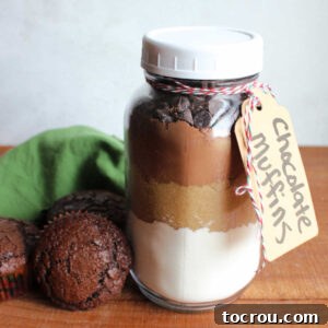 Jar of chocolate muffin mix with layers of flour mixture, brown sugar, cocoa powder, and chocolate chips next to a couple of chocolate muffins baked from the mix.