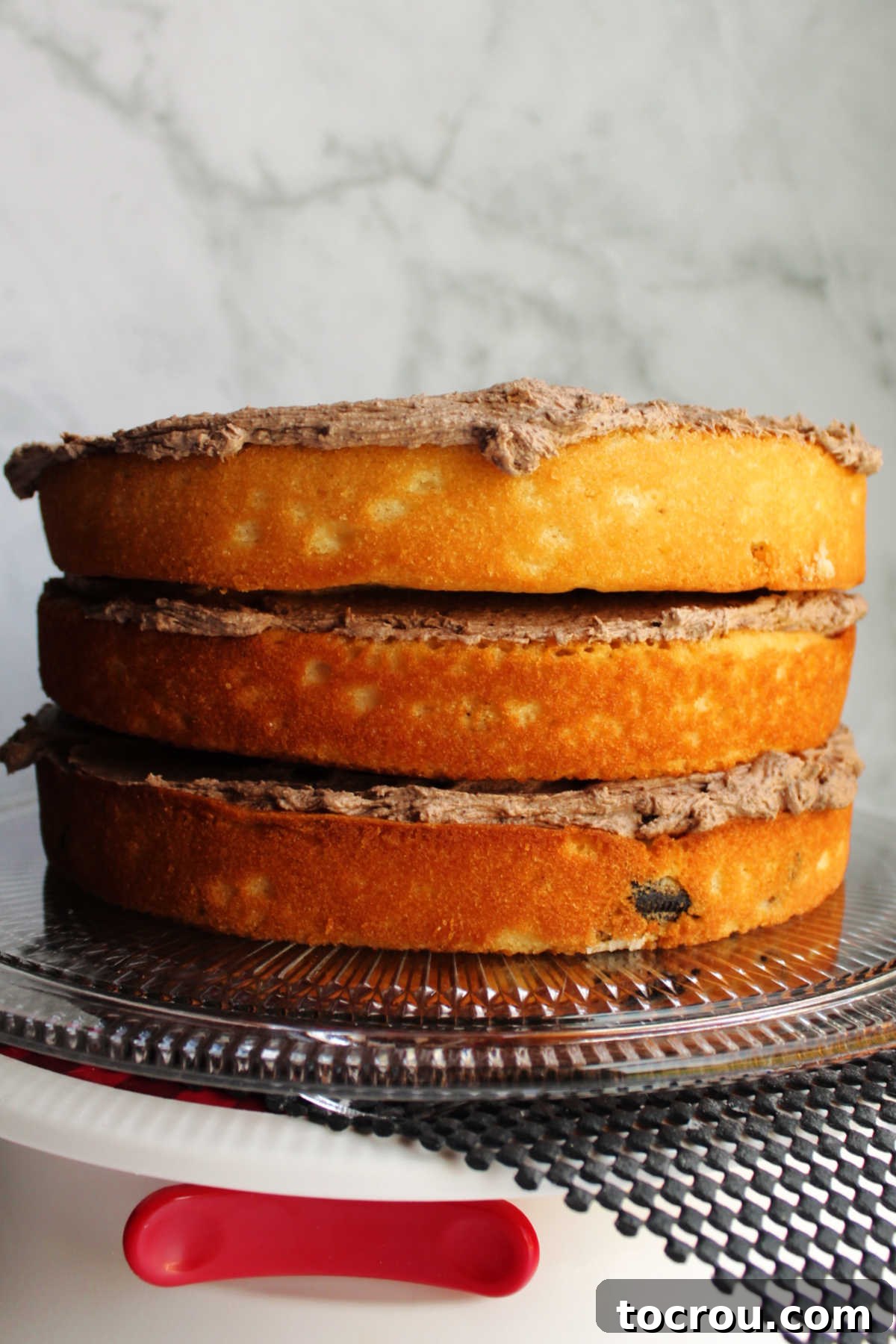 Building the layers of our delicious cookies and cream cake, one perfect layer at a time. Stacked layer cake with rich chocolate Oreo frosting spread between the layers, ready for the final outer frosting application.
