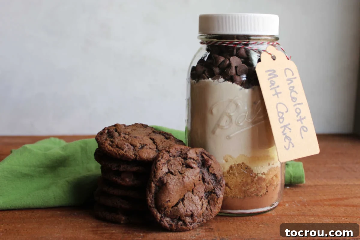 A quart jar filled with beautiful layers of dry ingredients for chocolate malt cookies sits beside a plate of freshly baked, super chocolaty looking chocolate malt cookies with visible chocolate chips. This image highlights the aesthetic appeal of the gift mix and the delicious end product.