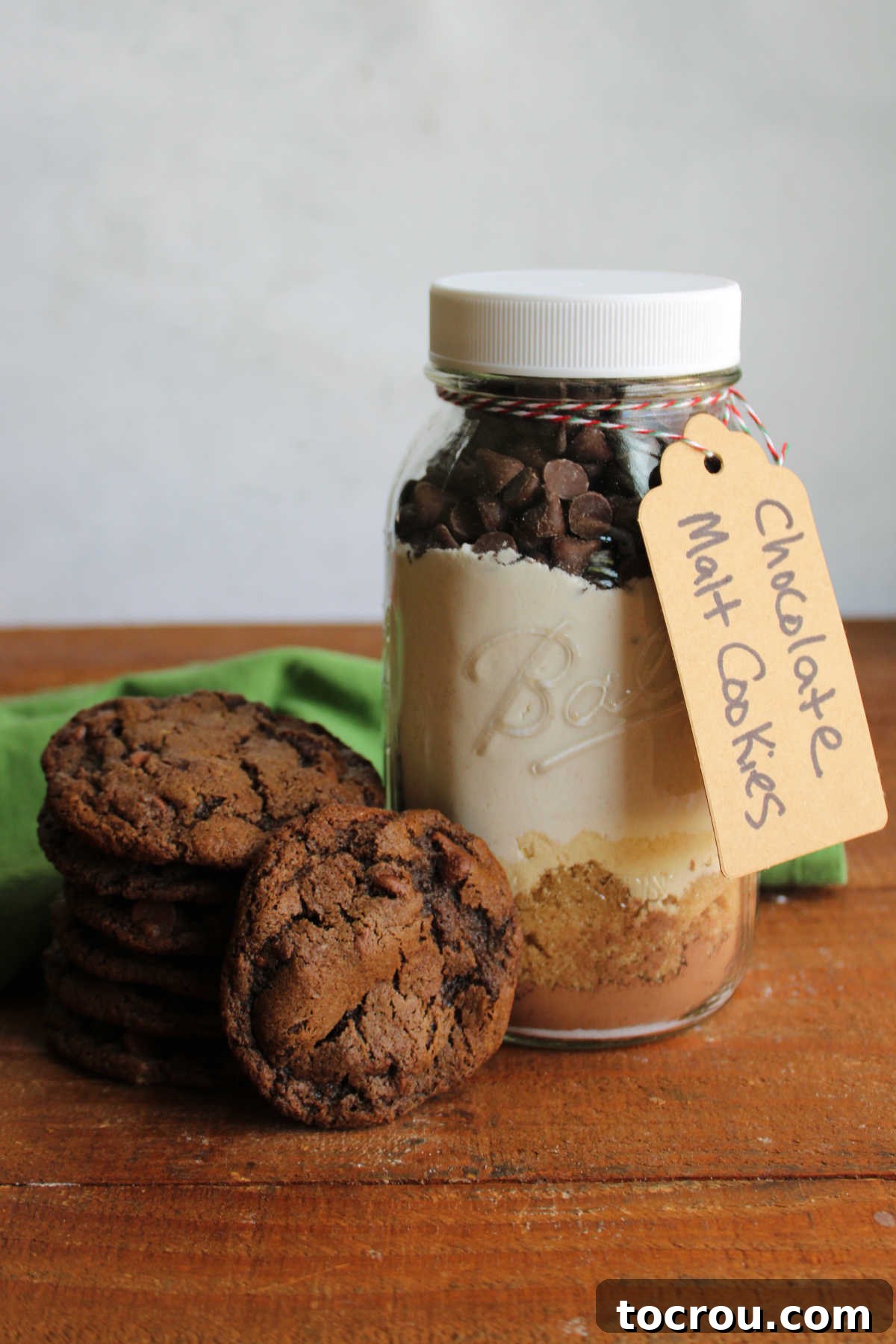 A stack of homemade malt chocolate cookies, generously studded with chocolate chips, rests next to a stylish jar of the dry mix used to create these delectable treats.