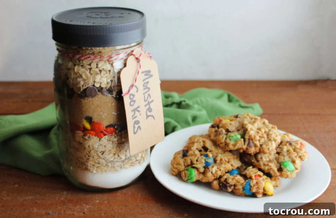 Jar of monster cookie mix with layers of sugar, oats, M&Ms, and chocolate chips next to a plate of monster cookies. 