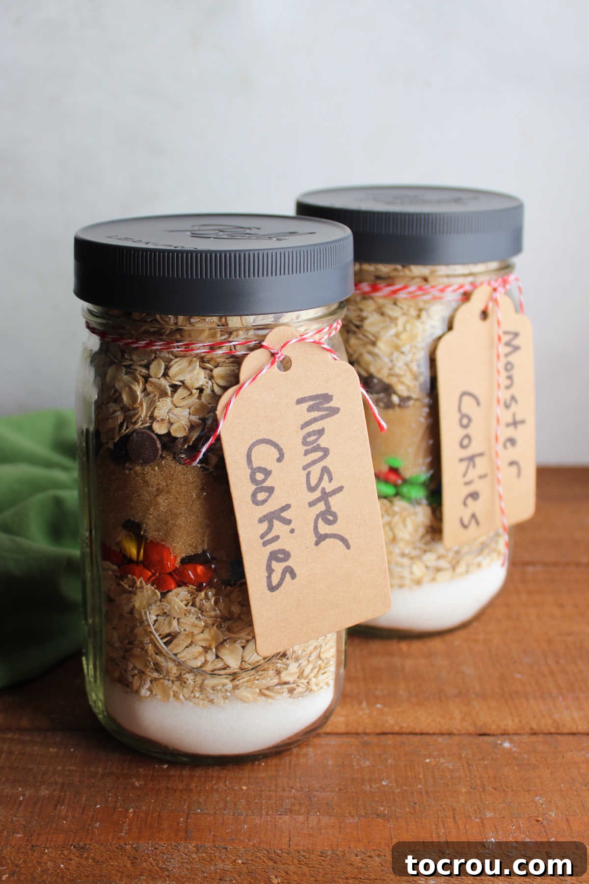 Jars of monster cookie mix with the dry ingredients in layers inside the jars and tags tied to the outside with baking instructions. 