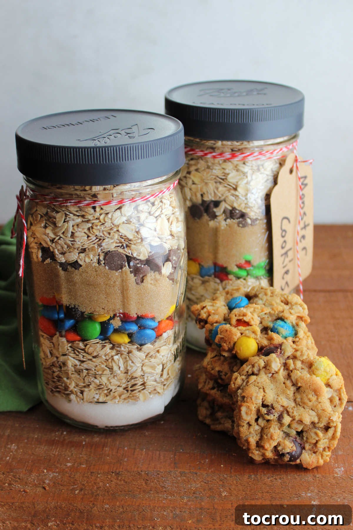 Two jars of homemade monster cookie mix next to a stack of homemade monster cookies. 