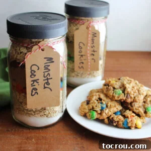 Two jars of layered monster cookie mix next to a plate of freshly baked monster cookies.