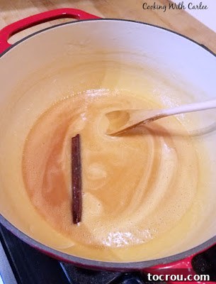 Cajeta Caramelizing on the Stove Enameled cast iron pot showcasing partially caramelized goat's milk with a cinnamon stick, mid-cooking process, thickening into a rich caramel.