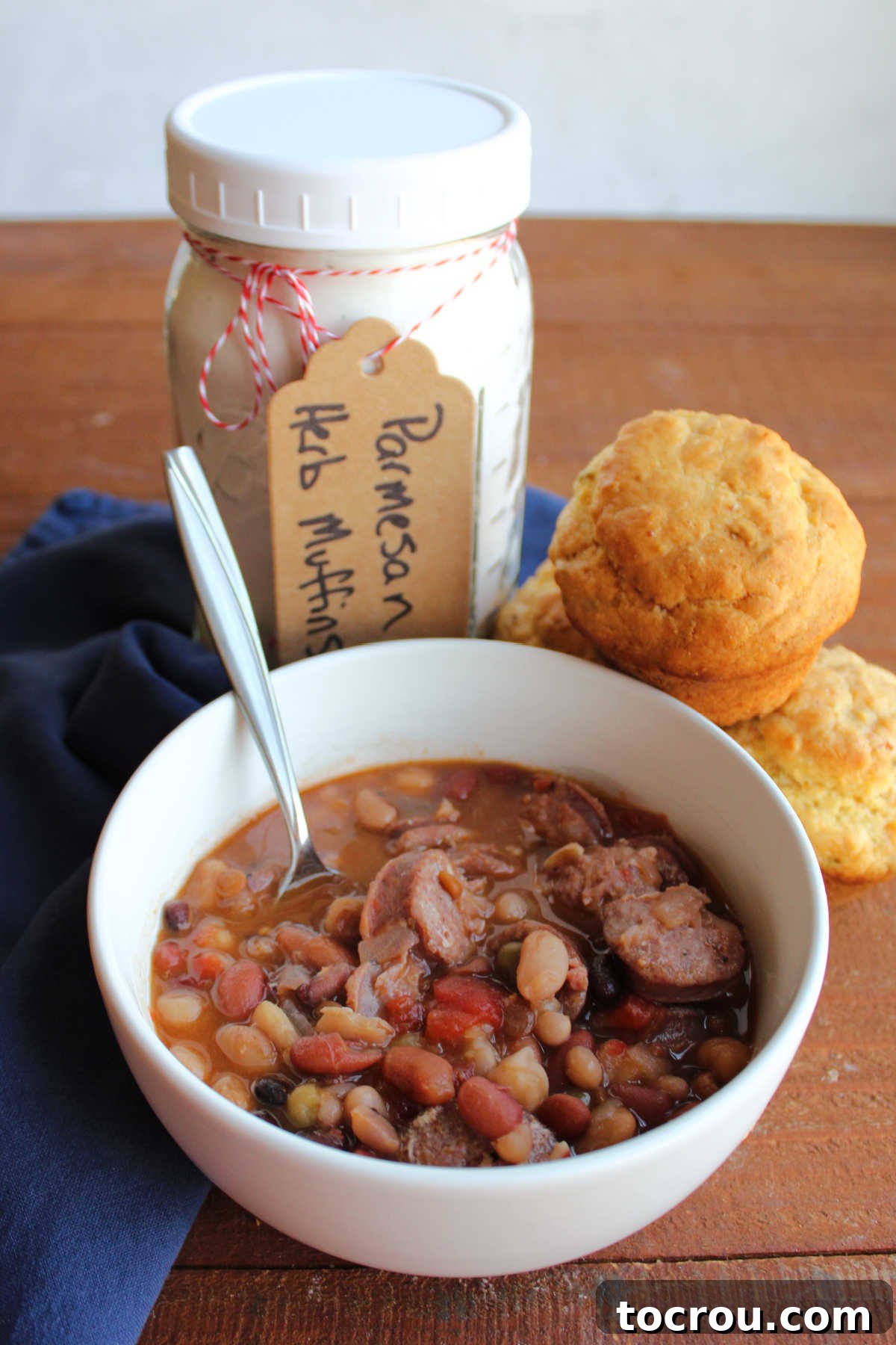 Jar of parmesan muffin mix and muffins next to a bowl of bean soup with sausage.
