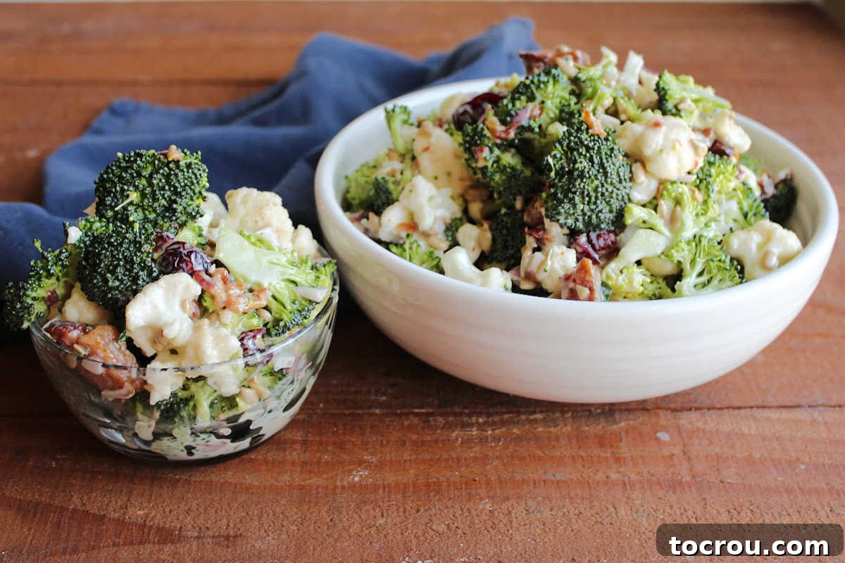 A small glass bowl with a serving of vibrant broccoli salad, placed next to a larger serving bowl brimming with more of this fresh side dish. It features crisp broccoli and cauliflower florets, golden sunflower seeds, ruby-red dried cranberries, and crumbled bacon, all coated in a creamy, luscious dressing. Fresh vegetables are artfully scattered around the bowls, suggesting garden-fresh ingredients.
